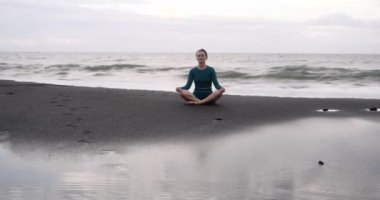 Lotus pozisyonunda genç bir kadın siyah bir kumsalda oturur ve büyük dalgalarla okyanusun arka planına karşı meditasyon yapar. Farkındalık ve zihnin saflığı. Konsantrasyon. Yaz sporları.