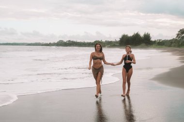 Çekici bir anne ve mayo giymiş genç kızı okyanusun yanındaki siyah kumsal boyunca koşuyorlar ve el ele tutuşup gülüyorlar. Yaz tatilinde çocuklu bir tatil. Anneler Günü.