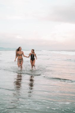 Mayolu iki tatlı kız kardeş sahilde okyanusun yanında koşup gülüyorlar. Genç bir kız ablasının elini tutar ve gülümser. Ailenle yaz tatilinde mutlu zaman geçir..