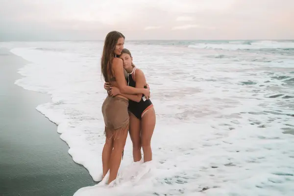 Büyük dalgalar ve şefkatle kucaklaşmalarla anne ve genç kızın plajda, okyanusun arka planında durduğu pankart. Bir çocuğa duyulan sevgi. Anneler Günü. Kızımla yaz tatili..