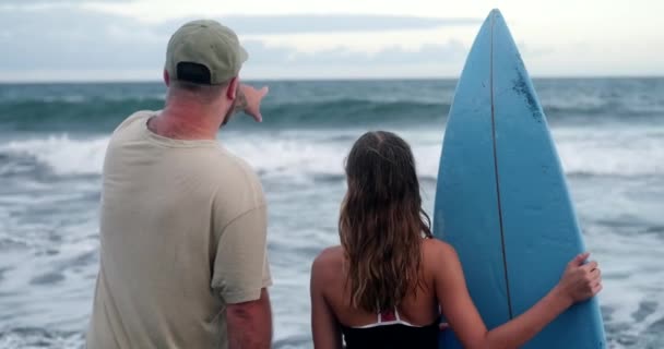 父亲和女儿面对大海站着 一个少女手里拿着一块冲浪板 她的父亲用手指着浪花 告诉她冲浪的原理 初学者冲浪 父亲节 — 图库视频影像