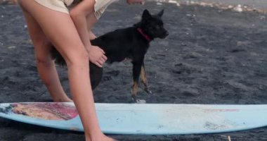 Küçük siyah köpeğine sörf tahtasında durmayı öğreten genç bir kızın yakın çekimi. Çocuk yaz tatilinde sahilde eğleniyor ve köpekle oynuyor. Tasasız çocukluk.