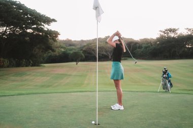 Spor etekli genç beyaz bir kadın bir golf kulübünün yeşil çimlerinde pratik yapan acemi bir golfçü. Kadın deliği ıskaladı ve üzgün, elinde sopayla başını tutuyor.