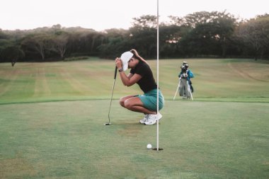 Acemi bir golfçü olan genç bir kadın golf sahasında çömeliyor ve elinde bir sopa tutuyor. O, deliği kaçırdığı ve rekabeti kaybettiği için üzgün. Yenilgiden dolayı hayal kırıklığı.