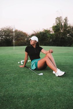 Genç çekici kadın profesyonel golf koçu yeşil çimlerde oturuyor ve poz veriyor. Golf kulübünde golf dersleri. Spor üniformalı bir öğretmenin portresi. Lüks yaz sporu.