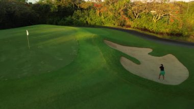 Profesyonel golf oynayan genç bir kadının kumdan bir golf sopasına vuruşunu yukarıdan aşağıya gösteren hava görüntüsü. Top tepede uçuyor, kadın deliğe çarpıp çarpmadığını görmek için zıplıyor..