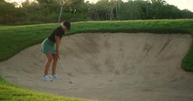 Genç bir bayan profesyonel golfçü kum tuzağına düştü. Topa tepedeki sığınaktan vuruyor ve çukura çarpıp çarpmadığına bakmak için yerden yukarı zıplıyor. Golf oynamayı zorlaştırıyor..