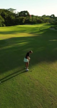 Yeşil bir çimenlikte golf oynayan genç bir kadının dikey hava görüntüsü. Sopasıyla topa sertçe vuruyor, çimenleri yerinden söküyor ve top uzaklığa doğru uçuyor..