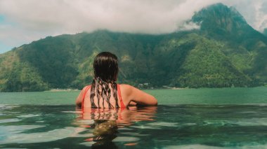 Tatilde kırmızı mayo giyen beyaz tenli genç bir kadın dağlara ve göle bakan sonsuz havuzlu bir tatil köyünde dinleniyor. Vücut için yaz tatilleri ve sağlık tedavileri.
