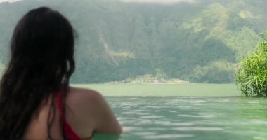 Yaz tatilinde olan genç bir kadın dağlık bir bölgedeki sıcak termal kaynaklarda dinleniyor. Havuzda oturuyor ve önündeki tepelere ve göle doğru bakıyor..