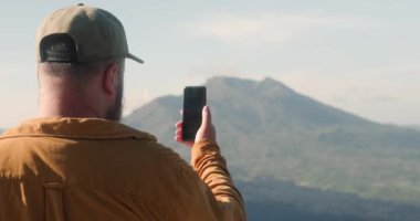 Bali 'deki dağlarda seyahat ederken volkan görüntüsünü telefonuyla çeken sakallı bir seyahat blogcusu. Bir blogcu macerası hakkındaki video hikayesini kaydeder.