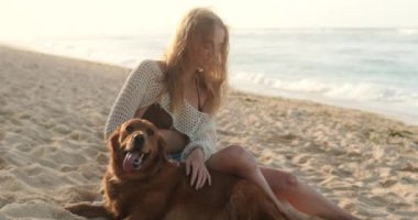 Çilli genç kızıl saçlı bir kadın okyanusun yakınındaki kumlu bir sahilde dilini dışarı çıkaran bir golden retriever ile oturuyor. Bir kadın nazikçe köpeğini okşar ve gülümser. Gün doğumunda yürümek.