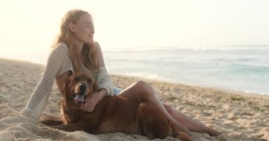 Genç, kızıl saçlı bir kadın okyanusun kıyısındaki kumlu bir sahilde Golden Retriever 'la oturuyor ve gülümsüyor. Bir kadın nazikçe köpeğini okşar ve ona sarılır. Sabahın köründe evcil hayvanınla ıssız bir sahilde yürüyorsun..