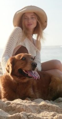Plajda Golden Retriever köpeğinin yanında oturan kızıl saçlı genç bir kadının dikey portresi. Köpeği okşar ve gülümser. Tropik bir adada evcil hayvanınla yaz tatili.