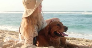 Genç ve çekici kızıl saçlı bir kadının portresi plajda oturur ve köpeğini mavi okyanusun arka planında okşar. Köpeğinle seyahat etmek ve tatilin tadını çıkarmak..