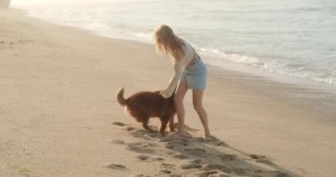 Genç kızıl saçlı bir kadın, sabahın ilk ışıklarıyla okyanus kıyısındaki plajda golden retriever köpeğiyle oynuyor. Köpek zıplar ve sahibinin ellerinden tasmayı kapar. Evcil bir hayvanla tatil.