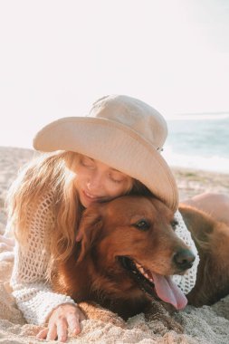 Kumlu sahilde çilli, çekici, kızıl saçlı bir kadının Golden Retriever 'a sarılışının dikey portresi. Kadınlar köpeklere, aşka ve şefkate gülümser ve sarılır. Yaz tatili yürüyüşü.