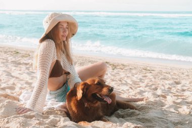 Genç, çekici, kızıl saçlı, şapkalı bir kadın kumsalda oturuyor ve köpeğini mavi okyanusun arka planında okşuyor. Köpeğinle seyahat etmek ve tatilin tadını çıkarmak..