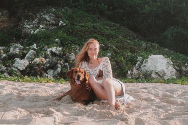 Genç, kızıl saçlı bir kadın okyanusun kıyısındaki kumlu bir sahilde Golden Retriever 'la oturuyor ve gülümsüyor. Bir kadın nazikçe köpeğini okşar ve ona sarılır. Sabahın köründe evcil hayvanınla ıssız bir sahilde yürüyorsun..