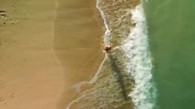 Bikini giymiş genç bir kadının turkuaz okyanus suyundan altın bir kumsala yürüyüşünün en üst görüntüsü. Yüzüyorsun ve yaz tatilinin tadını çıkarıyorsun. Kumda çıplak ayak izleri var..