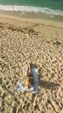 Hasır şapkalı genç kızıl saçlı bir kadının havadan görünüşü okyanus kıyısındaki beyaz bir battaniyede uzanıyor ve bir kitap okuyor. Yaz tatili boyunca dinlenme ortamı..