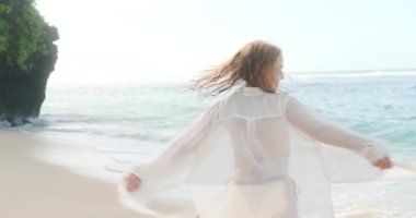 Kahverengi bikinili ve şeffaf beyaz tişörtlü neşeli genç bir kadın okyanus kenarındaki beyaz kumsalda dönüyor ve mutlulukla gülümsüyor. Uzun zamandır beklenen yaz tatili. Kumsalda bir sabah.