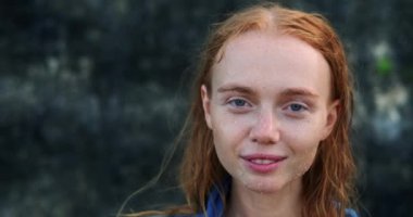 Mavi gömlekli, genç, kızıl saçlı bir kadın. Yüzünde çiller ve kum var. Okyanusun yanındaki iki kayanın arasında duruyor ve gülümsüyor. Yaz gezisi. Bir sırt çantasının portresi.