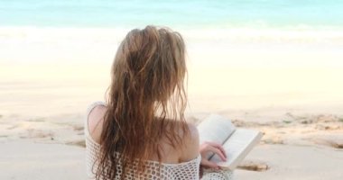 Mavi okyanus kıyısında, sahilde kitap okuyan, mayolu, kızıl saçlı genç bir kadının arka görüntüsü. Bir yaz tatilinin idealist bir resmi. Güneşte bronzlaşmak ve edebiyat okumak.