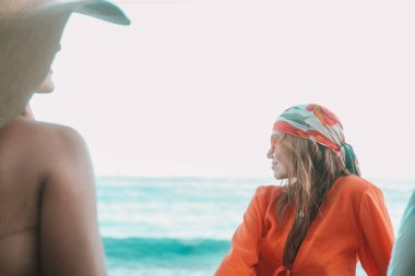 Tatil sırasında iki kız arkadaş sahilde oturuyor. Turuncu güneş gözlüklü kızıl saçlı bir kadın gülümseyip yüzünü çeviriyor. Şapkalı ikinci kadın sırtı kameraya dönük oturuyor. Arkadaşınla tatile çık.