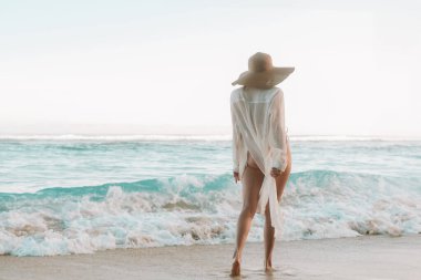 Beyaz şeffaf gömlekli, hasır şapkalı, mavi okyanusa ve dalgalara bakan kızıl saçlı genç bir kadının arka plan görüntüsü. Tropik bir adada yaz tatili konsepti.
