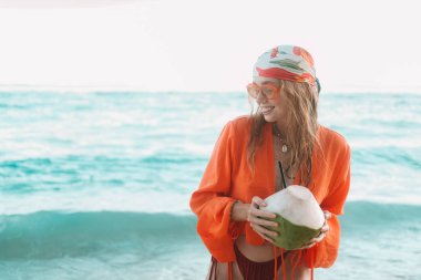 Turuncu güneş gözlüklü, mavi okyanusa karşı duran ve gülümseyen genç kızıl saçlı bir kadınla pankart. Elinde bir hindistan cevizi tutuyor. Yaz tatili, renkli bir adada.