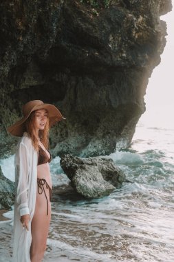 Hasır şapkalı, kahverengi bikinili ve beyaz şeffaf gömlekli genç kızıl saçlı bir kadın kayaların yanındaki sahilde duruyor, okyanus dalgaları kayalara çarpıyor ve su sıçrıyor. Yaz tatili.