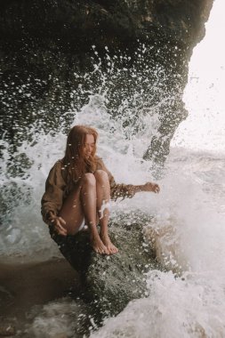 Bikinili genç kızıl saçlı bir kadın dev okyanus dalgalarının çarptığı bir kayanın üzerinde oturuyor, deniz köpüğü tepesine yükseliyor ve su sıçrıyor. Okyanusun arka planına karşı moda portresi. Özgürlük kavramı.