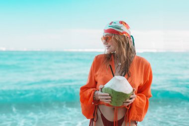 Turuncu güneş gözlüklü genç kızıl saçlı bir kadın ve mavi okyanusun karşısında duran bir gömlek. Elinde bir hindistan cevizi tutuyor. Tropikal bir adada renkli bir yaz tatili. Tatil