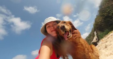 Plajda kırmızı mayo giymiş genç bir kadın Golden Retriever köpeğiyle selfie çekiyor. Köpeğe sarılıyor ve onun yüzünü öpüyor. Yaz tatili boyunca çadır ve köpekle yürüyüş yapmak..