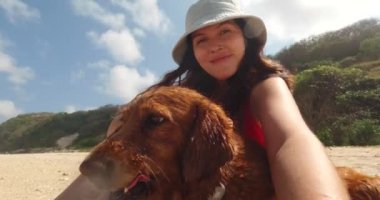 Genç bir kadın sahilde oturur ve av köpeğiyle aksiyon kamerasında selfie çeker. Arka planda bir adam çadır kuruyor. Yaz tatili boyunca aile kamp gezisi.