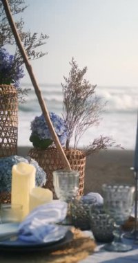 Okyanus yakınlarındaki siyah bir kumsal üzerindeki bir piknik masasının dikey görüntüsü, mavi bir masa örtüsüyle kaplıdır ve tabak ve bardaklarla servis edilir. Masada mumlar ve çiçekler var. Romantik.