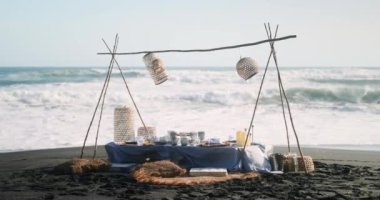 Okyanusun arka planına karşı siyah kumda büyük dalgalarla süslenmiş piknik alanı. Piknik mavi tonlarda, masada bir masa örtüsü ve tabaklarla dolu bir masa var. Romantik yemek..