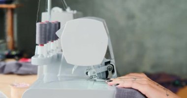 Dikiş makinesi olan bir masada oturan genç bir kadın terzinin elleri, gri pamuk kumaşa dikiş atıyor. Ev stüdyosu, küçük işletme. Elbise dikmek için..