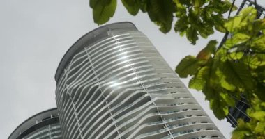 Yansıtıcı panellerle kaplı yüksek bir gökdelenin alt görüntüsü, önünde rüzgarda esen bir ağaç dalının yapraklarından oluşan bir kare var. Doğa ve modern binalar.