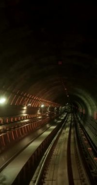 Yavaş çekimde dikey, bir metro tünelinin içinden geçen bir görüntüsü. Karanlık yuvarlak tünel parlak fenerler ve yer altındaki kıvrımlarla aydınlatılır. Büyük bir şehirde metro. Aktarım yolu.