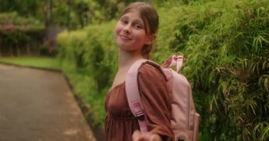 Omuzlarında pembe sırt çantasıyla genç bir kız okula koşmadan önce kameraya el sallıyor ve gülümsüyor. Okula dönelim. Yaz tatilinden sonraki ilk gün.