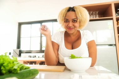 Mutfakta kıvrımlı, yeşil salata yiyen ve gülümseyen Afro-Amerikan kadın. Sağlıklı yaşam tarzı ve diyet konsepti. Artı boy kadın, vücut pozitifliği..