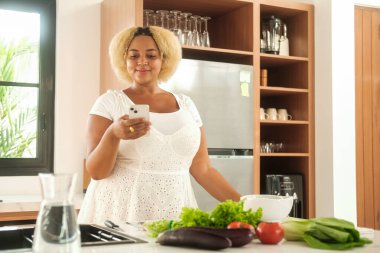 Afrikalı Amerikalı kıvrımlı kadın, elinde telefon ve sosyal medyada parşömenlerle parlak bir mutfakta duruyor. Taze sebzeler onun önündeki masada. Ayrıca blogcu salata hazırlıyor..