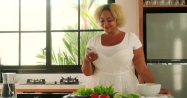 Afrikalı Amerikalı kıvrımlı kadın, elinde telefon ve sosyal medyada parşömenlerle parlak bir mutfakta duruyor. Taze sebzeler onun önündeki masada. Ayrıca blogcu salata hazırlıyor..