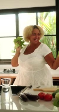 Yemek pişirirken mutfakta dans eden dikey Afro-Amerikan kadın. Masadan marul yapraklarını alıyor ve marakas gibi sallıyor. Çekici ve neşeli, iri yarı bir kadın. Vücut pozitif.