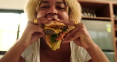 Afro-Amerikan kıvrımlı genç bir kadının portresi. Mutfakta etli ve peynirli büyük, sulu bir burger yiyor. Abur cubur ve sağlıksız yemek. Vücut pozitif.
