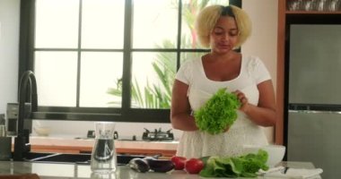 Genç Afrikalı Amerikalı kıvrımlı kadın mutfakta duruyor yeşil marul yapraklarından bir salata hazırlıyor, masada farklı sebzeler. Diyet ve sağlıklı yaşam tarzı. Vücut pozitif.