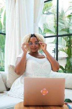 Oturma odasındaki kanepede oturup dizüstü bilgisayarla çalışan, gözlüklü, Afrikalı, kıvrımlı genç bir kadının portresi. Arka planda yeşil ağaçlar olan büyük pencereler. Uzak çalışma konsepti.