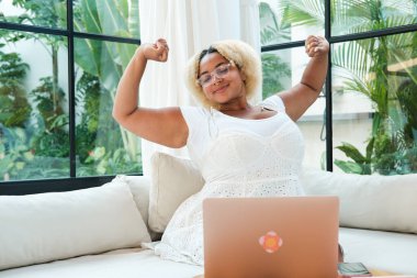 Oturma odasında dizüstü bilgisayarla çalışan Afrikalı, kıvrımlı, serbest çalışan kadın, günün sonunda yorgun ve tembelce esniyor. Uzaktan çalışma konsepti. Uykulu durum iş başında.